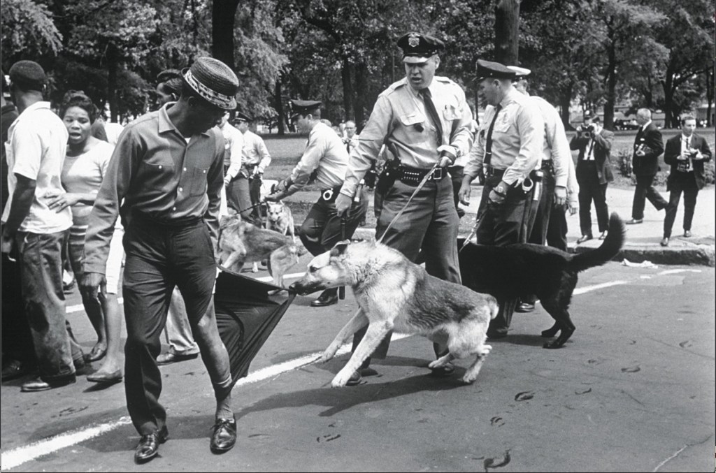 Two police officers encourage a dog to rip the pants off a Black man walking in Birmingham, AL.
