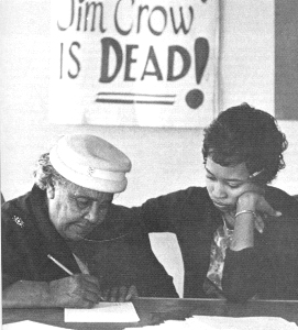 One woman instructs another woman who is writing on a paper. Behind them a sign reads "Jim Crow is dead!"
