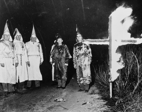 Five men in Ku Klux Klan robes and pointy hats burn a cross in NC in 1965.
