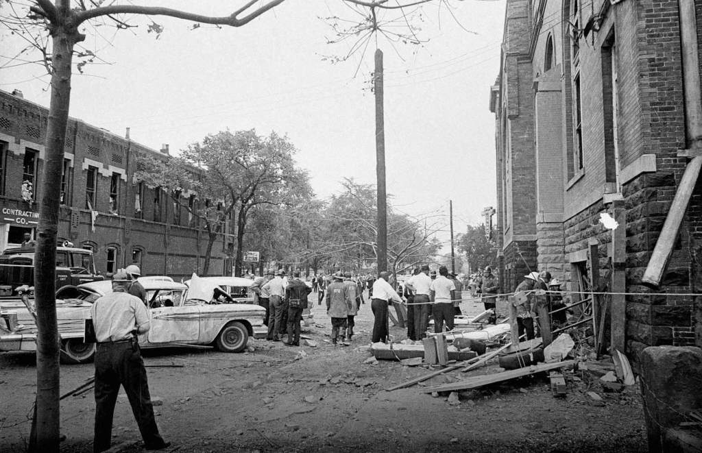 A crowd including police gather around debris from the bombed Sixteenth Street Baptist Church.
