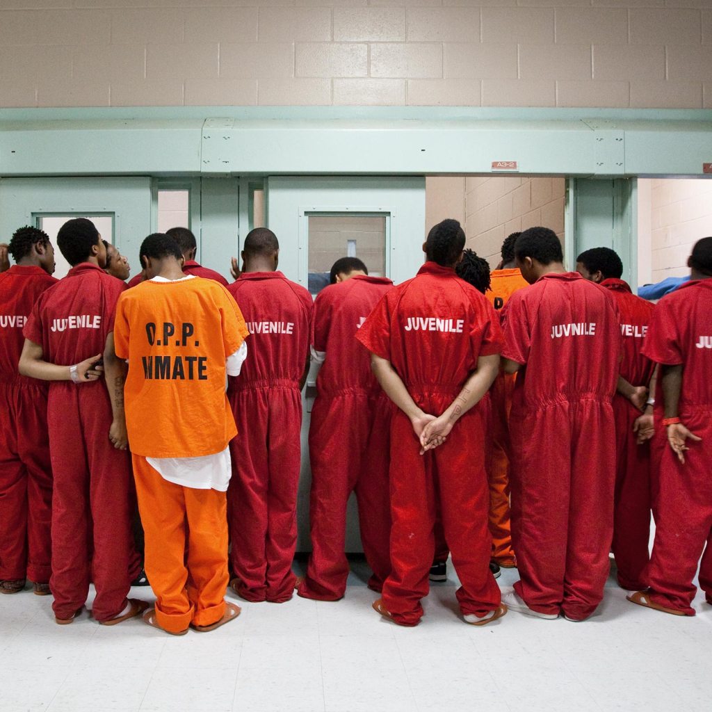 Photo of the backs of 14 African American inmates wearing uniforms saying "Juvenile."